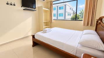 A hotel room with a bed, a TV mounted on the wall, a window with curtains, and a view of buildings outside.