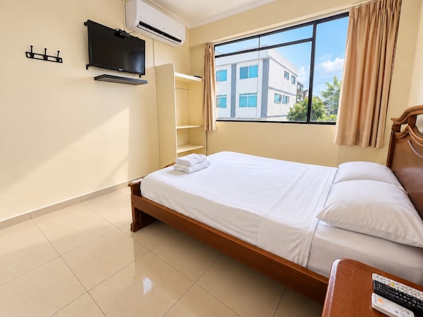 A hotel room with a bed, a TV mounted on the wall, a window with curtains, and a view of buildings outside.