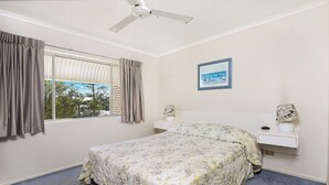 Quarto com cama, ventoinha no teto, janela com cortinas, mesa de cabeceira e quadro na parede.