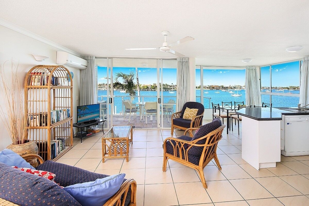 A spacious living area with a balcony overlooking a marina, furnished with wicker chairs and a sofa, and a small kitchen area.