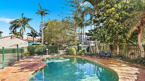 Uma piscina com borda curva, cercada por um alpendre de tijolos e uma cerca verde.