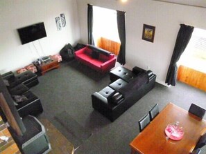 A living room with a black sofa set, a red couch, a wooden dining table with chairs, and a TV mounted on the wall. - Mackenzie Lodge (Twizel)