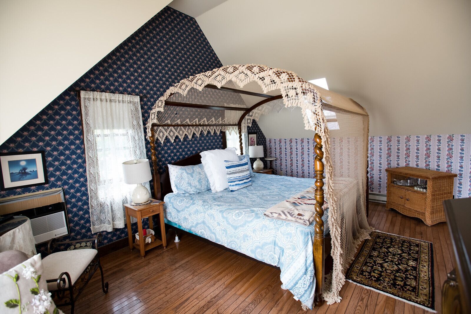 A bedroom with a four-poster bed, a wooden nightstand, a bedside table, a wicker chest of drawers, and a patterned rug.