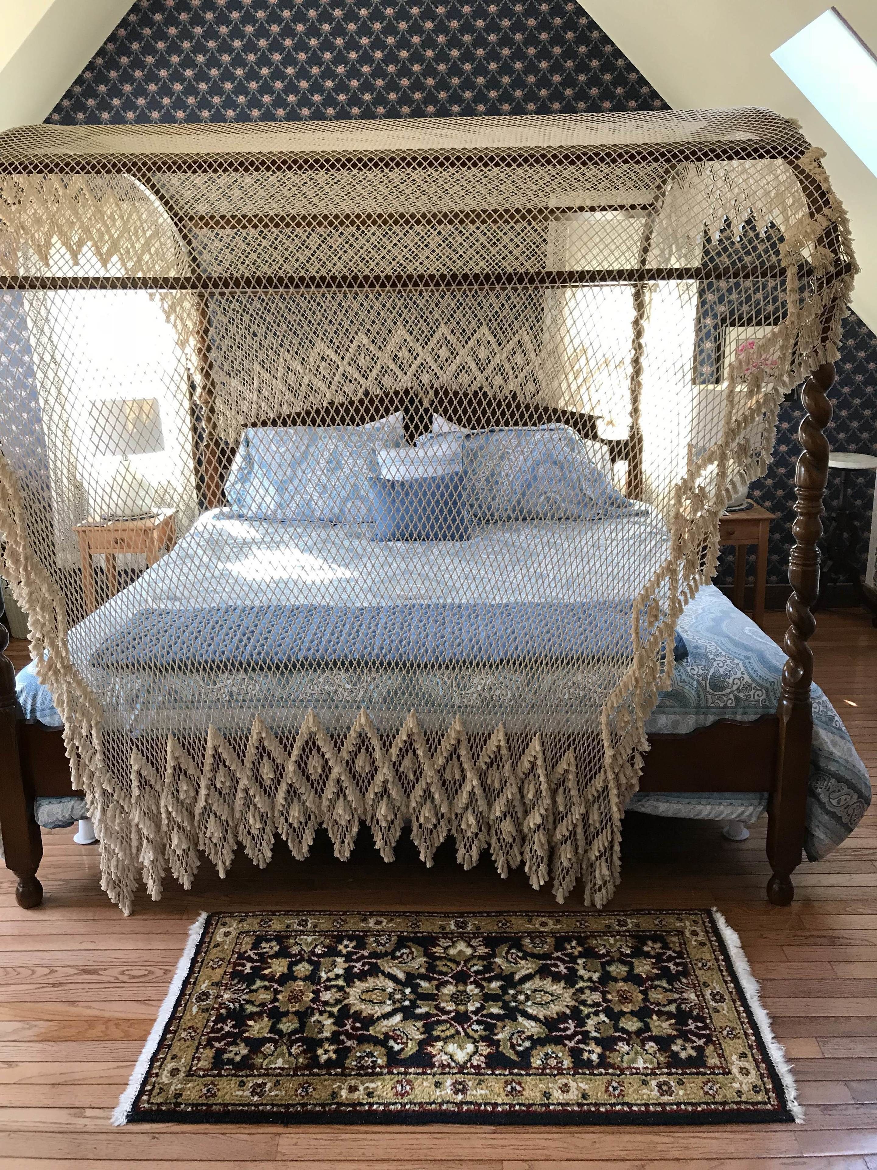 Un lit à baldaquin à quatre poteaux, surmonté d’un dais en dentelle, agrémenté d’une couvre-lit à motifs et d’un tapis foncé et décoratif posé sur un plancher en bois.