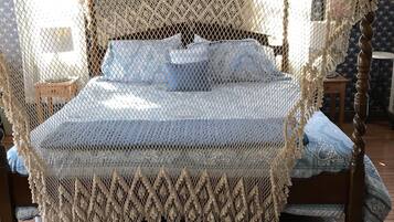 Un lit à baldaquin à quatre poteaux, surmonté d’un dais en dentelle, agrémenté d’une couvre-lit à motifs et d’un tapis foncé et décoratif posé sur un plancher en bois.