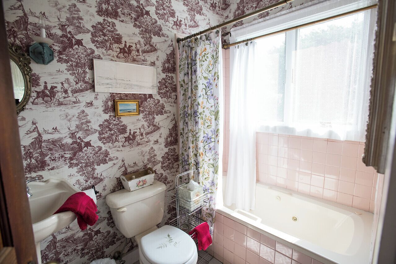 Une salle de bain comprenant des toilettes, un lavabo et une baignoire. On y trouve une fenêtre avec des rideaux, un miroir et une étagère fixée au mur où sont rangés les articles de toilette.