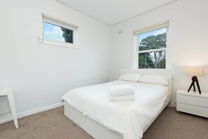 A bedroom with a bed, two windows, a bedside table, and a lamp.