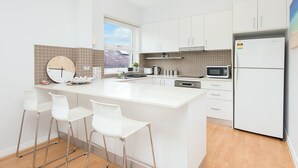 A modern kitchen with white cabinets, a large island with bar stools, a built-in microwave, and a refrigerator. - Central Bondi Apartment New H321 (Bondi Junction)