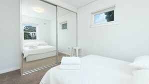 A bedroom with a large mirror, a bed with white linens, a small window, and a white nightstand. - Central Bondi Apartment New H321 (Bondi Junction)