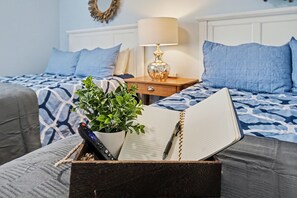 Ein ordentlich bezogenes Bett mit einer gemusterten Tagesdecke, eine Nachttischlampe, ein Topf mit Pflanze und ein offenes Notizbuch mit Stift auf einer Holzkiste.