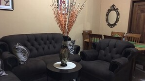 A living room with a dark-colored sofa, a small round table with a vase, and a dining area with wooden chairs and a mirror.