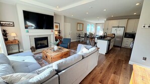 Amplia sala de estar con chimenea, un televisor grande, una cocina con armarios blancos y un comedor.