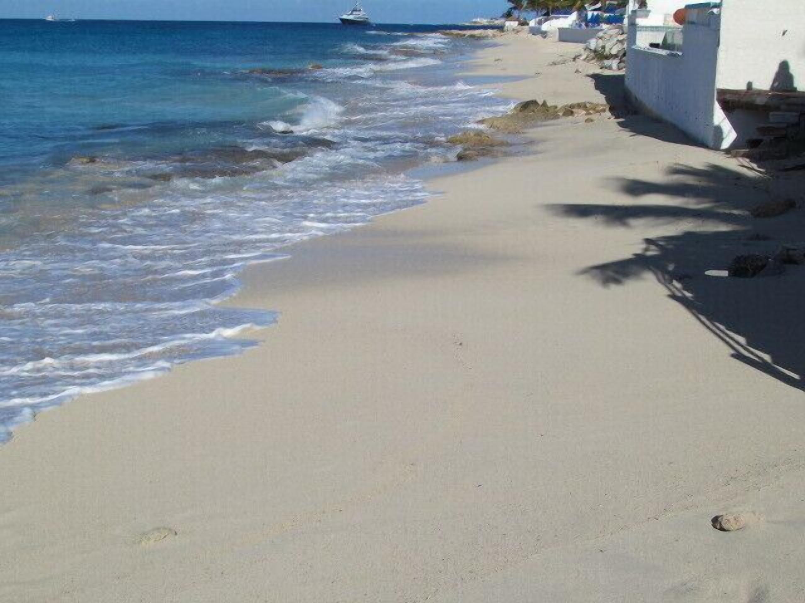 Una playa con un edificio blanco, cielo despejado y un barco a lo lejos.