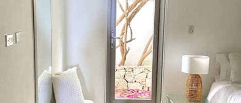 Une chambre moderne avec un banc, une table en verre surmontée d’une lampe, et une grande fenêtre donnant sur un mur de pierre et les branches d’un arbre.