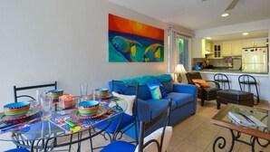 A living room with a blue sofa, a dining table with colorful bowls, and a kitchen in the background. - Restful Oceanfront 1BR, Northshore,  Low Density building with only 12 condos. (Cozumel)