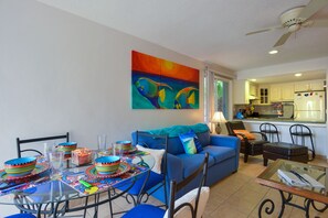 A living room with a blue sofa, a dining table with colorful bowls, and a kitchen in the background.