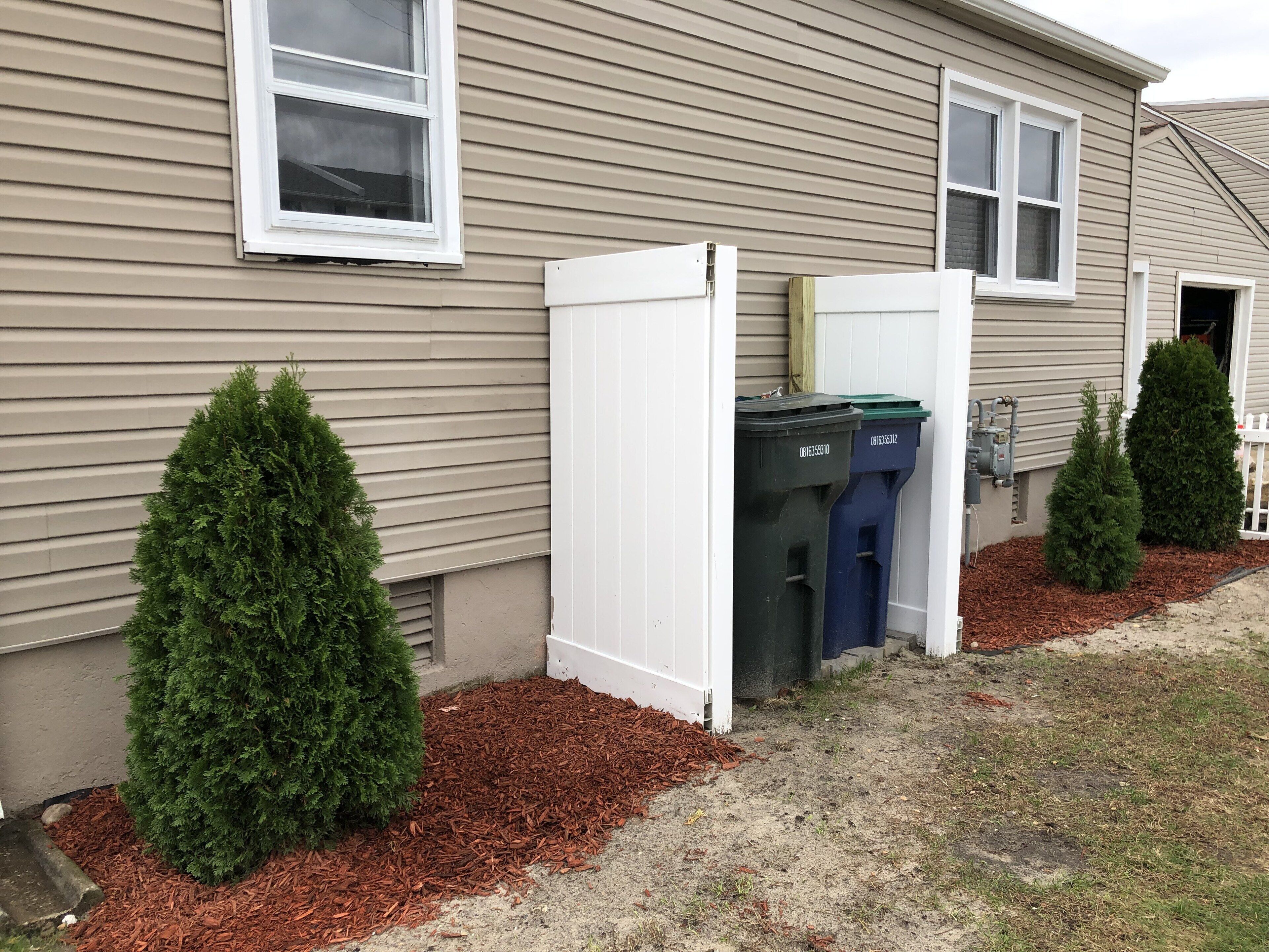 Un immeuble résidentiel à la façade beige, doté de deux fenêtres et d’une porte blanche. On y trouve deux arbustes verts et deux poubelles avec des étiquettes indiquant qu’elles sont réservées au recyclage.