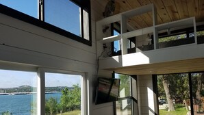 A room with a large window overlooking a body of water, a TV mounted on the wall, and a sofa.