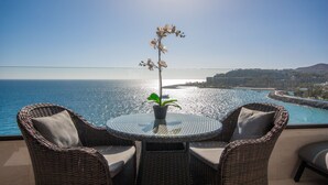 A round table with two wicker chairs and a potted orchid, overlooking a coastal view with a clear sky.