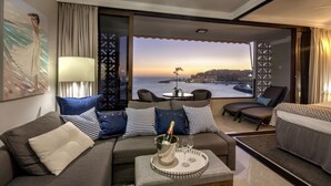 A hotel room with a sofa, armchairs, a dining table, and a view of the city at sunset.