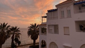 A coastal building with a balcony overlooking the sea, palm trees, and a sunset sky.
