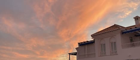A coastal building with a balcony overlooking the sea, palm trees, and a sunset sky.