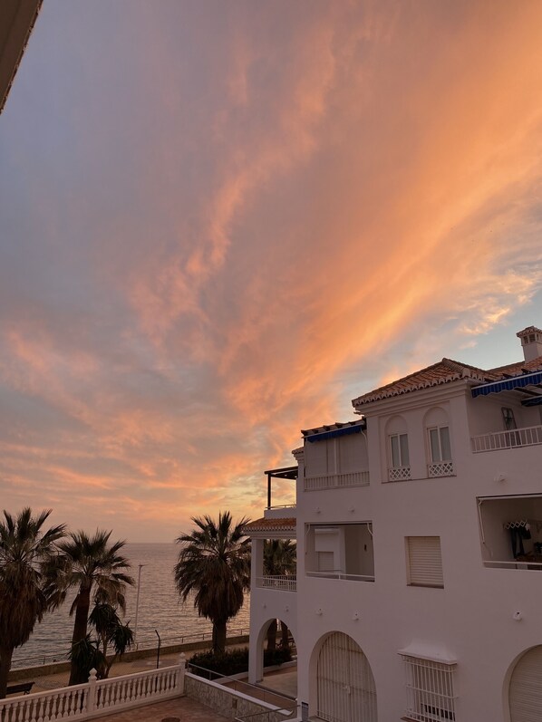 Ein Küstengebäude mit Balkon und Blick auf das Meer, Palmen und einem Sonnenuntergangshimmel.