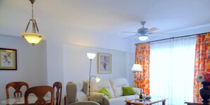 Ein Wohnzimmer mit einer Couch, Sesseln, einem Couchtisch, einer Stehlampe, einem Deckenventilator und Vorhängen.