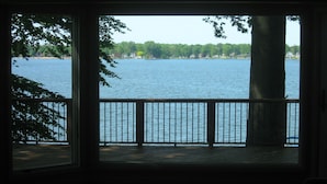 A view from a covered deck overlooking a lake with a wooden railing and a large tree in the center.