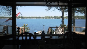A view from a covered deck overlooking a lake, with chairs and tables arranged for dining.