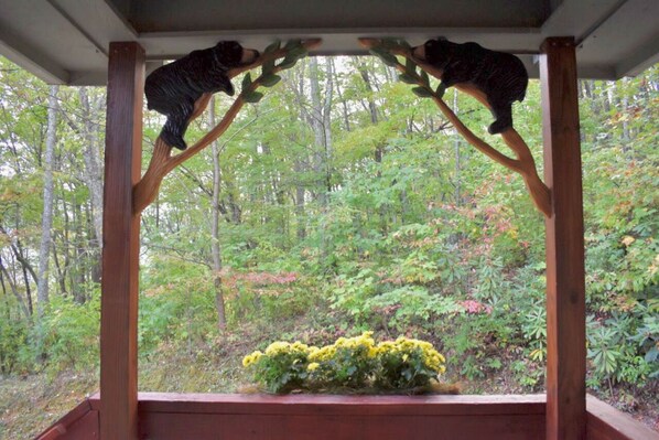 A wooden gazebo with two bear sculptures on branches, surrounded by lush greenery and a small yellow flower bed.