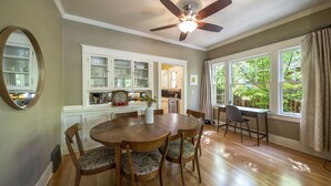 A dining area with a round wooden table, chairs, a ceiling fan, and a view of a kitchen and garden through large windows. - Historic Craftsman Bungalow, Just Steps to Downtown ~HOT TUB!~ (Bend)