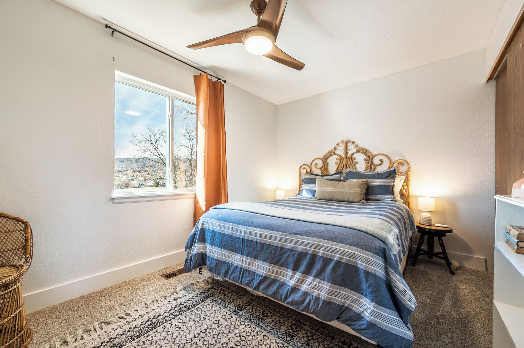 Ein Schlafzimmer mit einem großen Bett, einem Deckenventilator, einem Fenster mit Vorhängen, einem Nachttisch mit Lampe und einem Korbstuhl.
