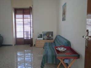 A living room with a sofa, a small table with a television and decorative items, a door with a window, and a tiled floor.