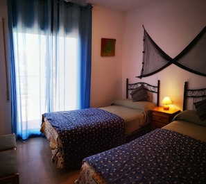 A room with two beds, a nightstand with a lamp, a chair, and a window with curtains.