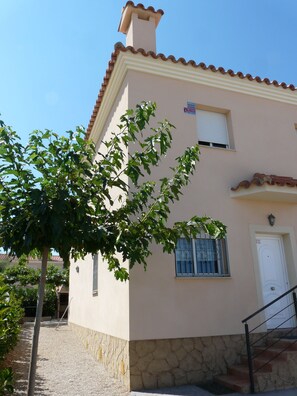 A two-story building with a stone base and a beige upper section. There is a small window with metal bars and a white door.
