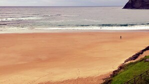 A beach with a person walking, a cliff in the background, and a cloudy sky. - Casa de Las Flores Apartment with private garden in front of the beach of Comillas (Comillas)