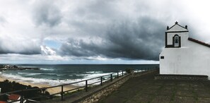 A coastal view with a white building featuring a bell tower, a stone wall, and a beach in the foreground. - Casa de Las Flores Apartment with private garden in front of the beach of Comillas (Comillas)