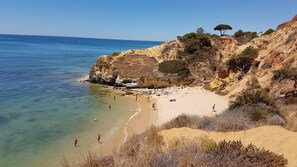 A coastal beach with clear waters, rocky cliffs, and sparse vegetation.