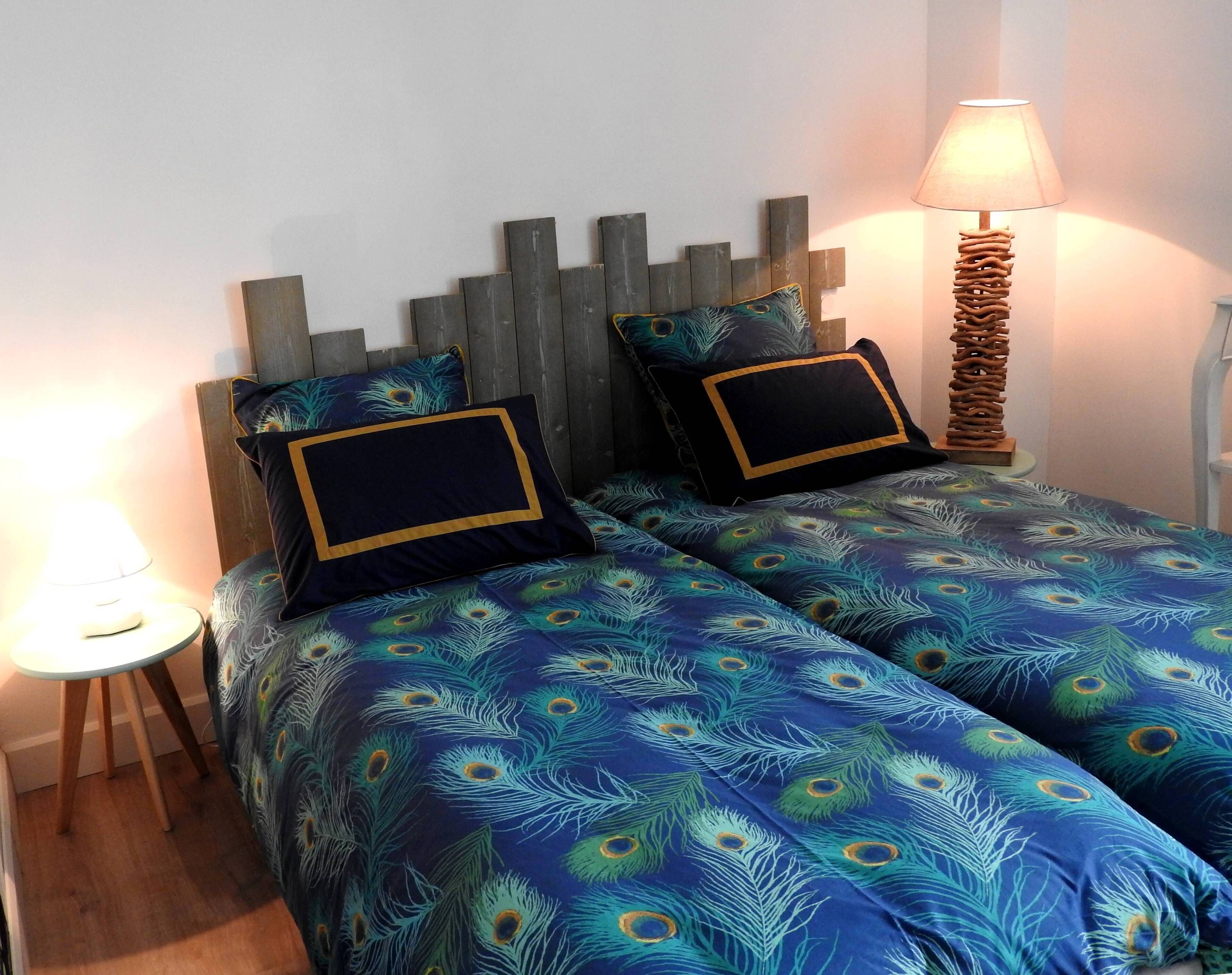 A bedroom with two beds, each with a wooden headboard and peacock feather patterned bedding. There is a bedside lamp on each side of the beds.