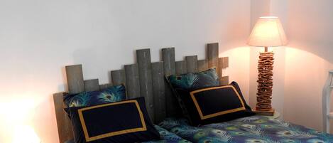A bedroom with two beds, each with a wooden headboard and peacock feather patterned bedding. There is a bedside lamp on each side of the beds.