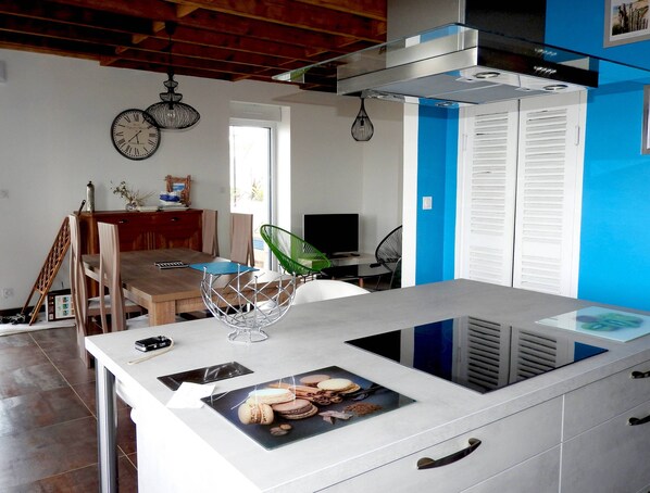 Una cocina moderna con una isla central, un comedor con sillas de madera, un televisor y un ventanal que proporciona luz natural.