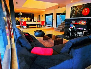Een moderne woonkamer met een grote zwarte bank, een rode sierkussen en een zwart voetenbankje. Er staat een salontafel en een televisie op een dressoir. De kamer heeft een groot raam met uitzicht op de stad.