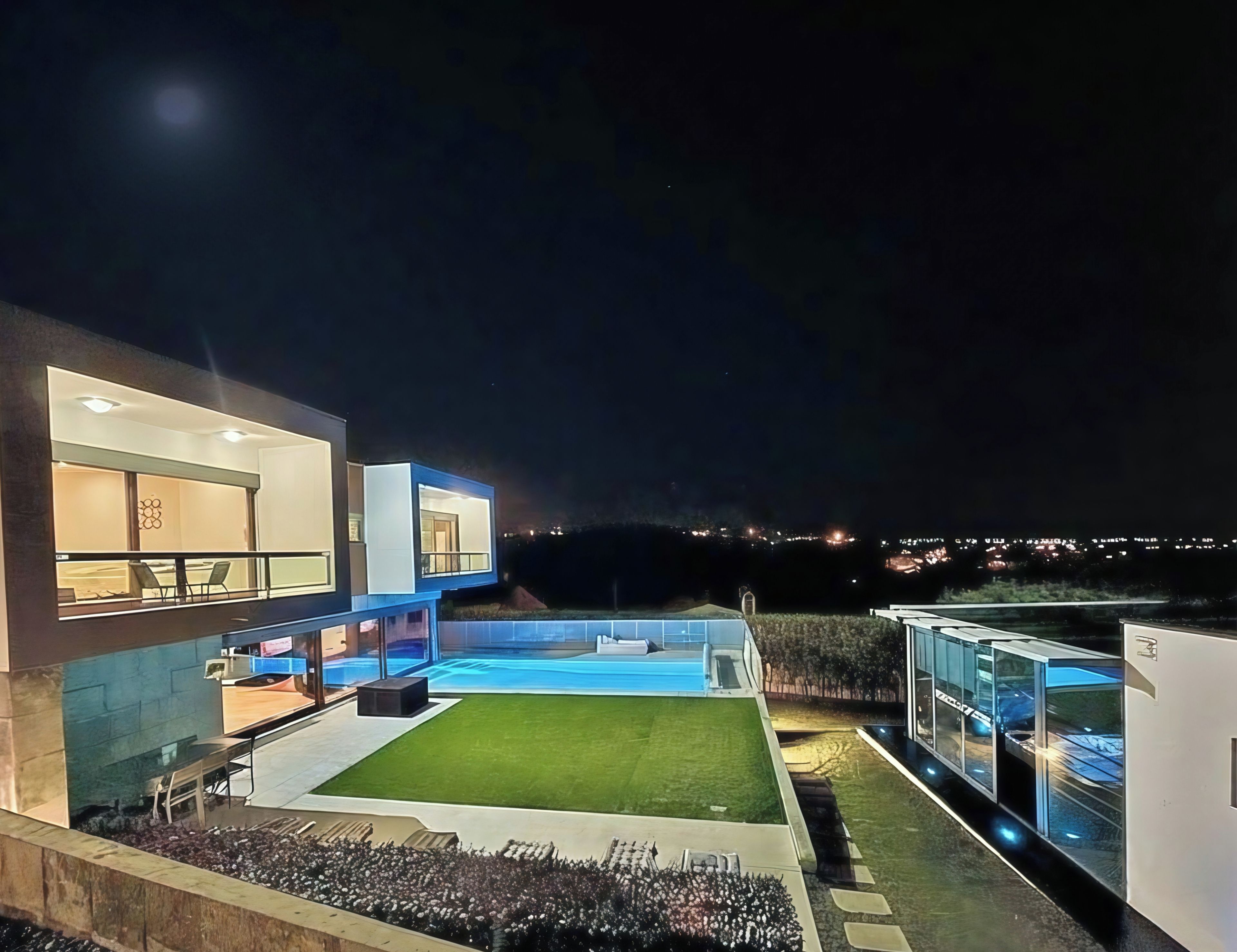 Een modern huis met een zwembad, een verzorgd gazon en uitzicht op de stad 's nachts.
