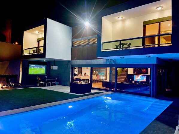 Una casa moderna de dos pisos con piscina, mobiliario exterior y televisor de pantalla plana.