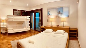 A hotel room with a bunk bed, a single bed, a wooden nightstand with lamps, a small table with a lamp, and a view of the beach through the glass door. - I L❤️VE U! Villa. Sea view! SPA! Heated Pool! Cinema! Private Chef! (Ericeira)