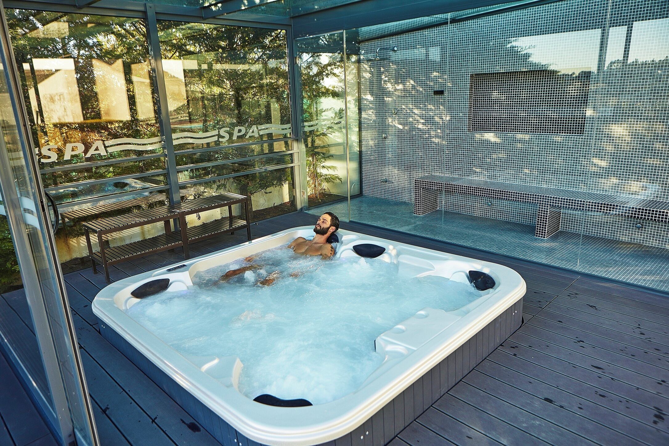 A hot tub with a person relaxing, surrounded by glass walls and a wooden deck.