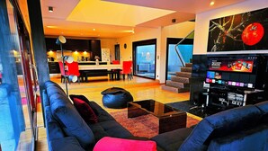 A modern living room with a black sofa, red cushions, a coffee table, and a television displaying channels. - I L❤️VE U! Villa. Sea view! SPA! Heated Pool! Cinema! Private Chef! (Ericeira)
