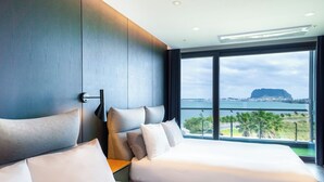 Ein modernes Hotelzimmer mit zwei Betten, einem großen Fenster mit Blick auf das Meer und eine entfernte Berglandschaft, sowie einem minimalistischen Design.