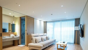 Ein modernes Hotelzimmer mit einer Couch, einem kleinen Tisch, einem an der Wand befestigten Fernseher, einem Badezimmer mit Dusche und einem großen Fenster mit Vorhängen.